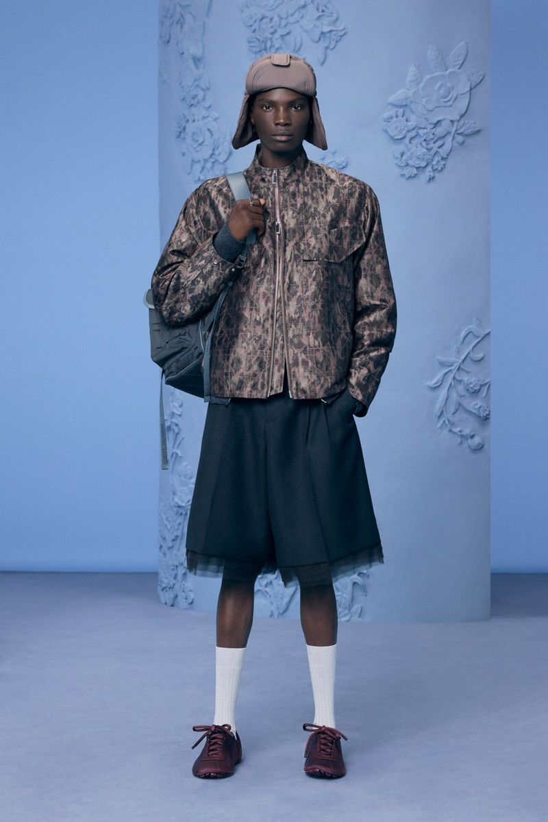 Model wearing a patterned jacket, shorts, and trapper hat against a blue studio background for Dior Men