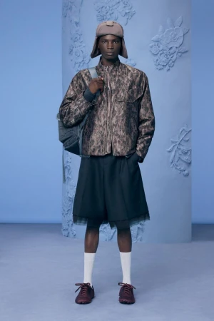 Model wearing a patterned jacket, shorts, and trapper hat against a blue studio background for Dior Men