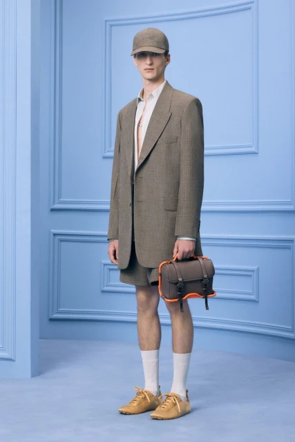 Full-length studio shot of a model wearing a brown suit with matching baseball cap, shorts, shoes, socks, and a small brown handbag.