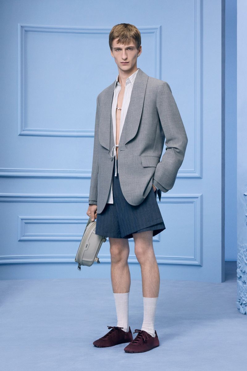 A slender man showcases a grey jacket, open shirt, dark shorts, and red shoes against a light blue panel wall in a studio setting.