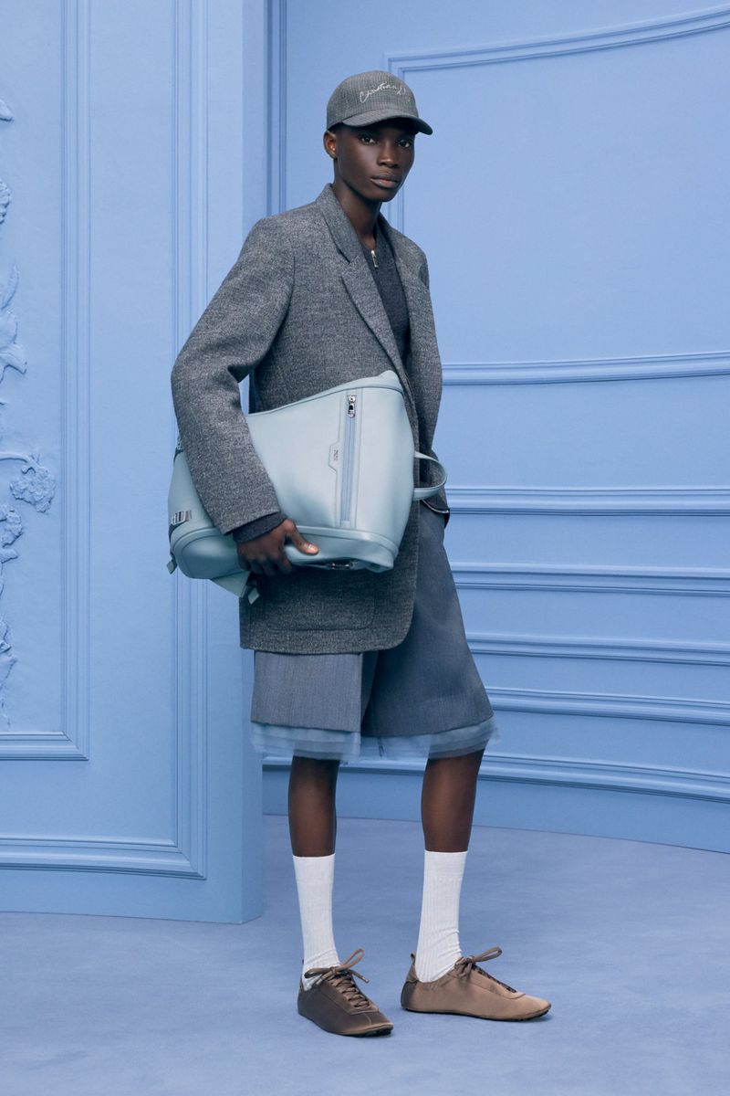 Fashionable person stands against a light blue wall holding a bag. They wear a blazer, shorts, cap, socks, and shoes in a studio setting.
