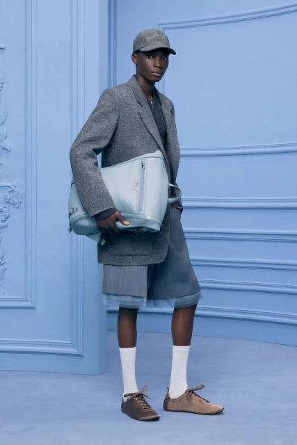 Fashionable person stands against a light blue wall holding a bag. They wear a blazer, shorts, cap, socks, and shoes in a studio setting.