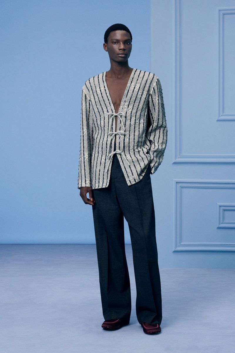 Elegant Black male model poses in a vertically striped sequined cardigan with a deep V-neck and dark wide-leg pants against a light blue backdrop.
