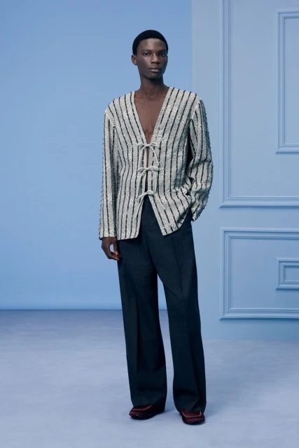 Elegant Black male model poses in a vertically striped sequined cardigan with a deep V-neck and dark wide-leg pants against a light blue backdrop.