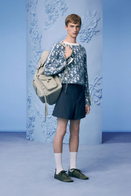 Fashion shot of a young male model wearing a Dior sweater, dark shorts, and sneakers, with a backpack in a studio setting.