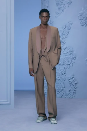 A male model wearing a brown suit jacket and matching trousers against a pale blue floral relief wall background.