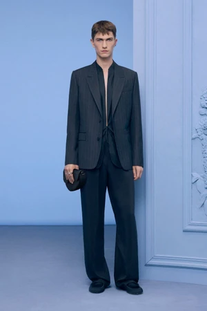 Model wearing a dark pinstripe suit and holding a black handbag against a light blue studio backdrop.