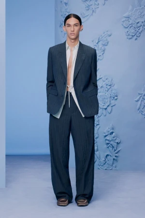Model wearing a tailored dark suit jacket and matching trousers against a light blue textured studio background