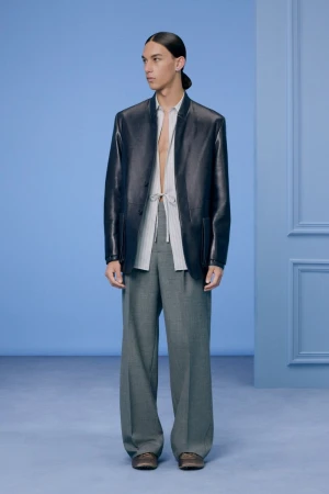 Model in dark leather jacket and grey trousers standing against a pale blue architectural backdrop for Dior Men Pre-Fall 2024.