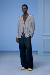 Model wearing a grey V-neck cardigan and dark trousers against a blue studio background for Dior Men Pre-Fall 2024