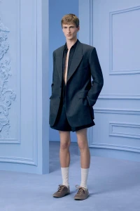 Male model wearing a dark oversized blazer and matching shorts with white socks and sneakers in a blue classical studio set.