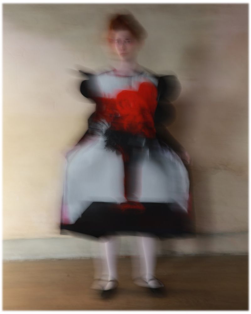 Young woman in a patterned dress stands indoors with motion blur. The dress features red, white, and black colors with an artistic design.