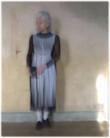 An elderly woman with white hair wearing a sleeveless grey dress over black sheer sleeves against a neutral wall.