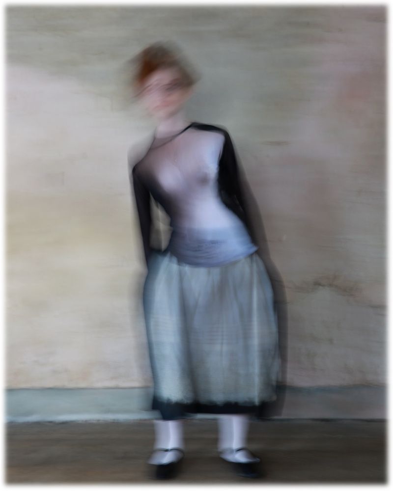 Full-length studio shot of a blurry, redhead adult female in a two-tone top and light skirt, posed against a light-colored wall with soft, diffused lighting.