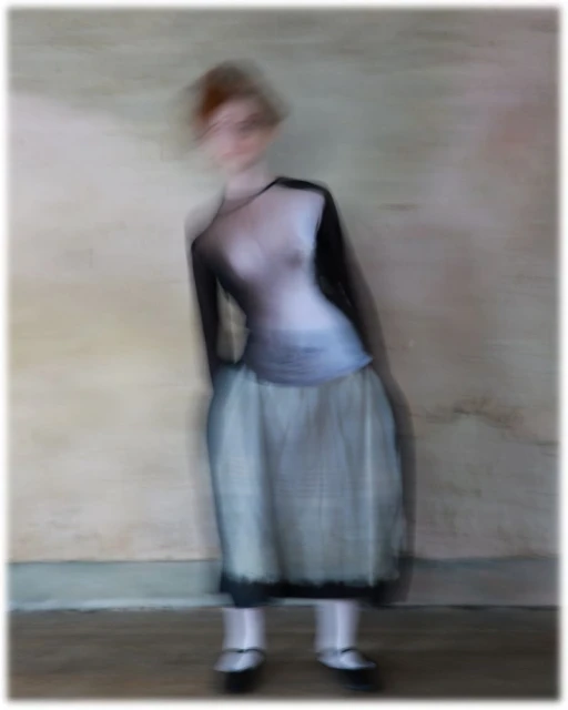 Full-length studio shot of a blurry, redhead adult female in a two-tone top and light skirt, posed against a light-colored wall with soft, diffused lighting.