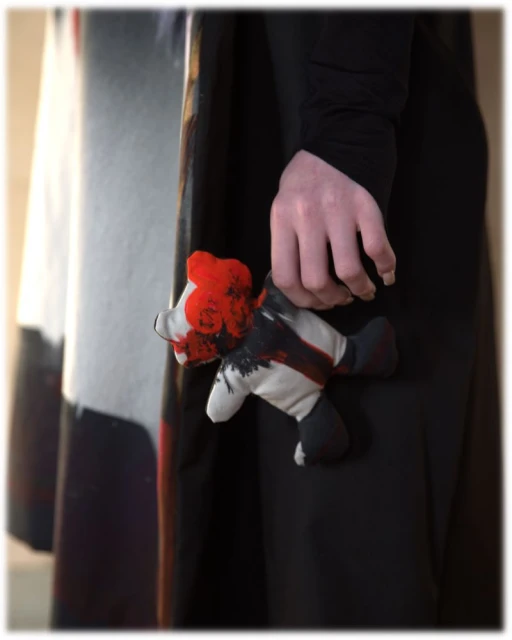 Close-up of a person's hand holding a handmade plush toy with black and red paint on a white background, showcasing texture and design.