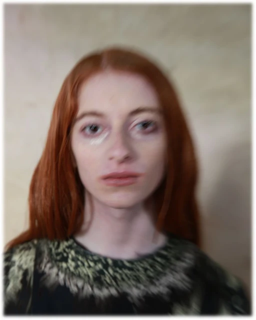 Portrait of a red-haired female adult with fair skin, wearing a dark top with light-colored designs, against a neutral blurred background.