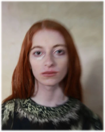 Portrait of a red-haired female adult with fair skin, wearing a dark top with light-colored designs, against a neutral blurred background.