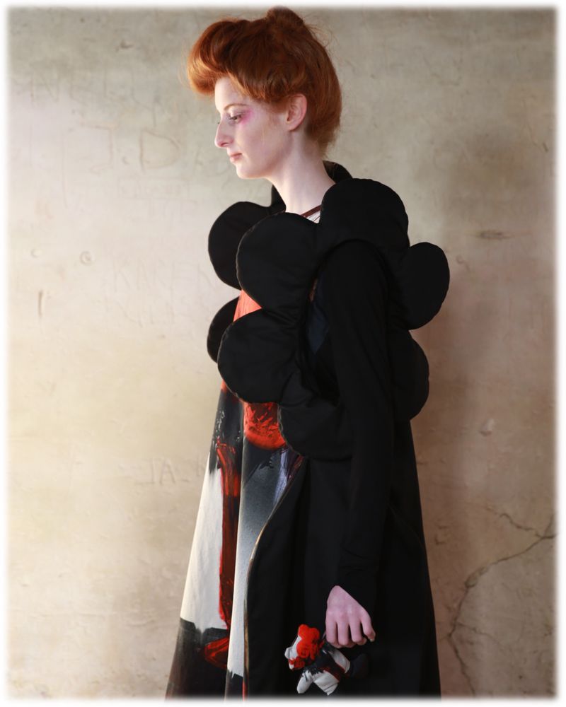 Woman in abstract black couture coat against textured wall. Art fashion portrait featuring model and hand-held doll in studio light.