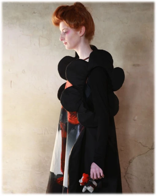 Woman in abstract black couture coat against textured wall. Art fashion portrait featuring model and hand-held doll in studio light.