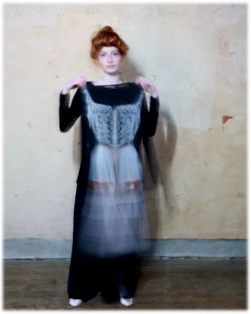 Fashion photo of a red-haired woman holding up a dress with a blurred effect, standing in an interior with neutral, textured walls.