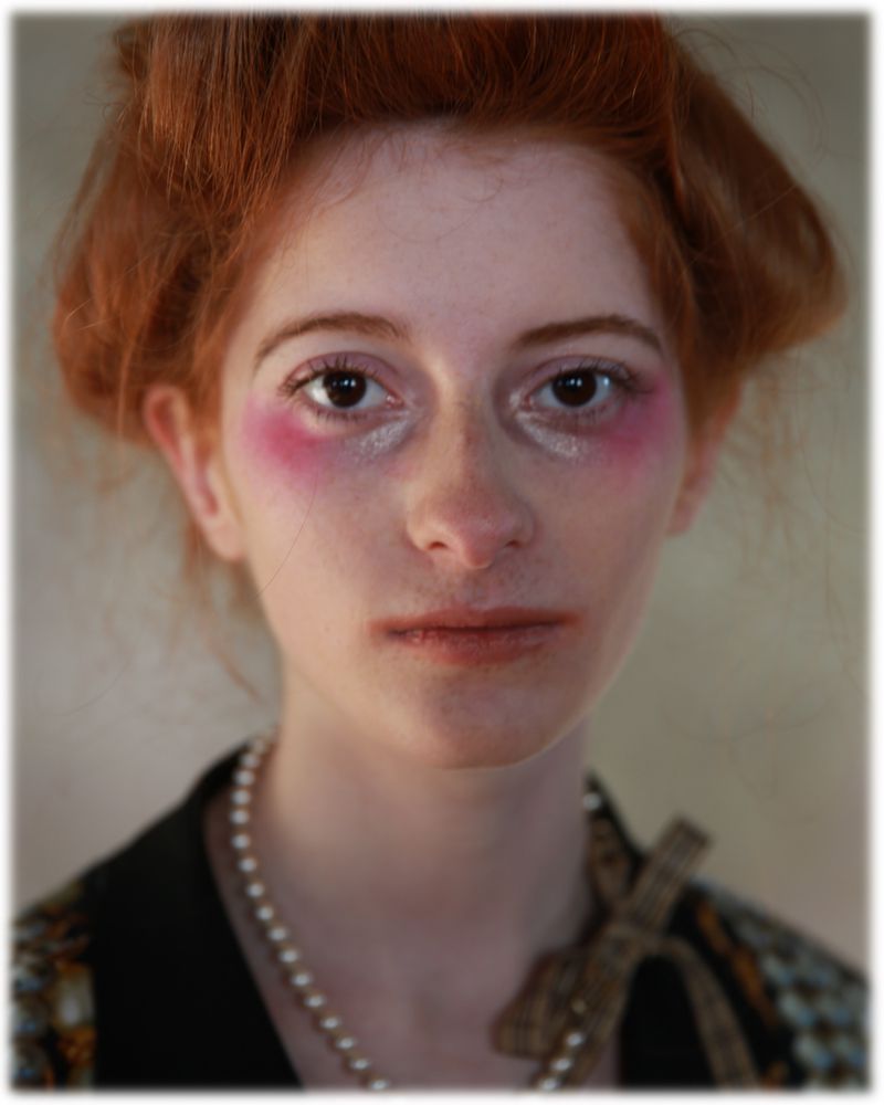Close-up portrait of a young woman with auburn hair, wearing pearl necklace, looking directly at camera with dramatic pink blush.