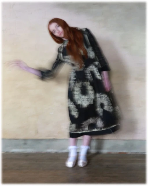 Full-length portrait of a woman with long red hair, wearing a black-and-white dress and white shoes, posing in front of a cream-colored wall.