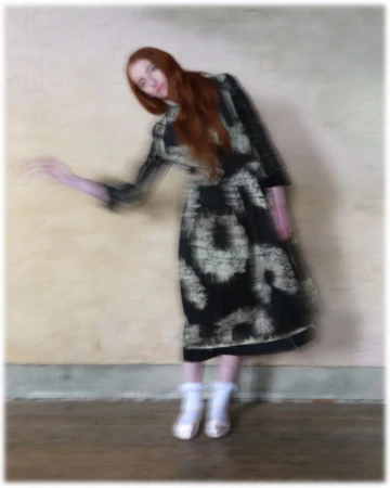 Full-length portrait of a woman with long red hair, wearing a black-and-white dress and white shoes, posing in front of a cream-colored wall.