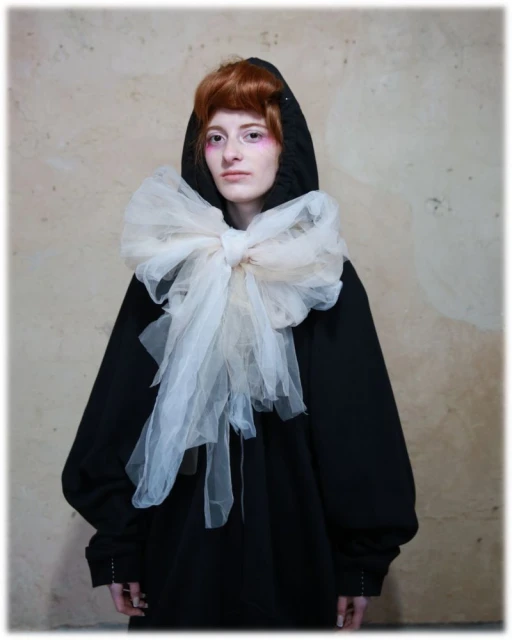 Portrait of a redhead woman wearing a black hooded sweatshirt and a large white tulle bow, posing in front of a textured wall.