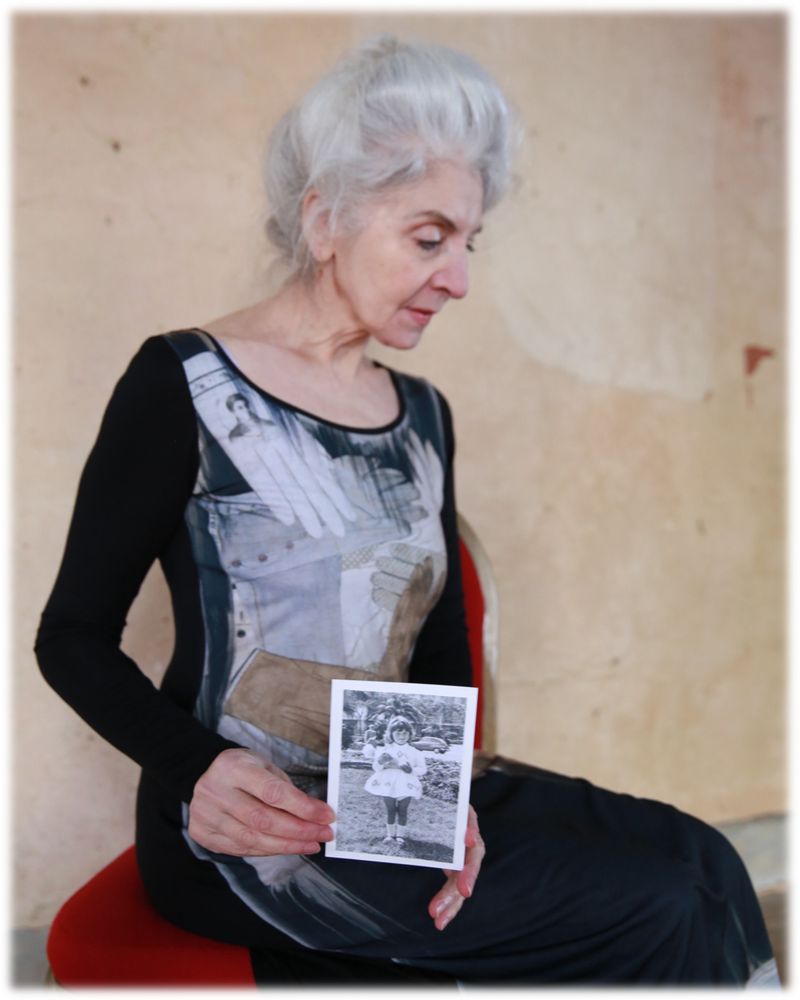 Elderly woman with gray hair holds a black and white photograph while seated indoors, wearing a black long-sleeved dress with a printed bodice.