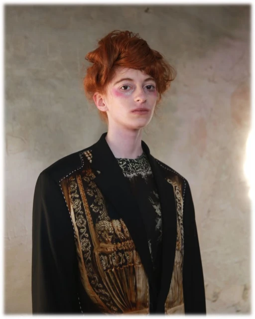 Portrait of a person with red hair wearing a black blazer with detailed gold embellishments, set against a textured beige backdrop.