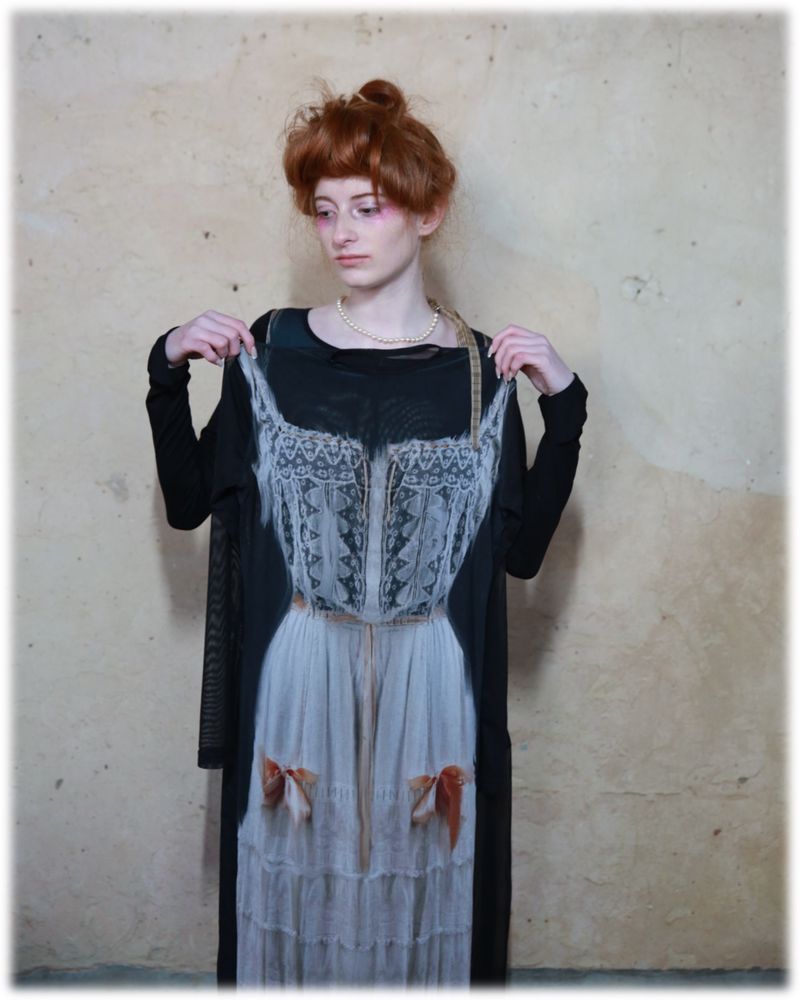 Portrait of a red-haired woman wearing a vintage-style black dress, lace top, and pearl necklace, holding a white lace corset.