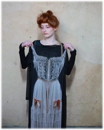 Model with red hair wearing a black dress holding up a printed slip garment against a mottled textured wall.