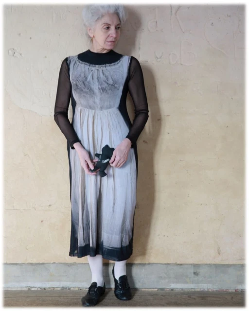 Mature woman in a grey ombre dress stands in front of a textured wall. She is holding a small stuffed animal toy. Neutral indoor light.