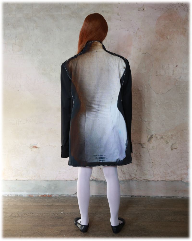 Back view of red-haired woman wearing oversized blazer dress with tailor's dummy print, white tights, and black ballet flats indoors.