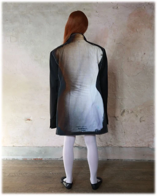 Back view of red-haired woman wearing oversized blazer dress with tailor's dummy print, white tights, and black ballet flats indoors.