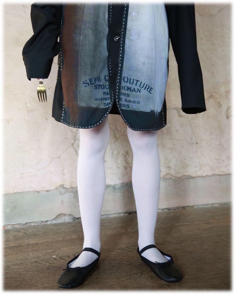 Child stands in front of a peeling wall wearing a black blazer and dress with white tights and black flats, holding a fork.