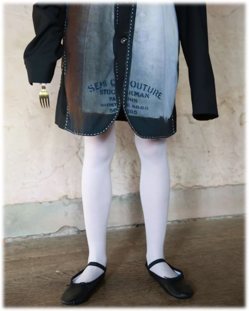 Child stands in front of a peeling wall wearing a black blazer and dress with white tights and black flats, holding a fork.