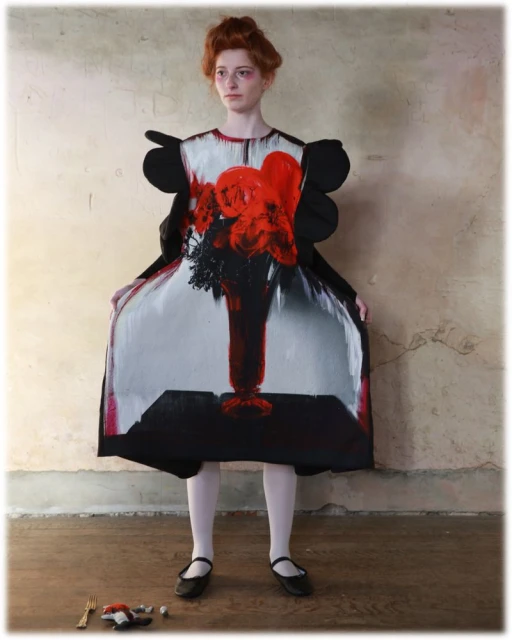 A redhead model in a studio setting poses in a dress featuring a vibrant vase of flowers print, complemented by white tights and flats.