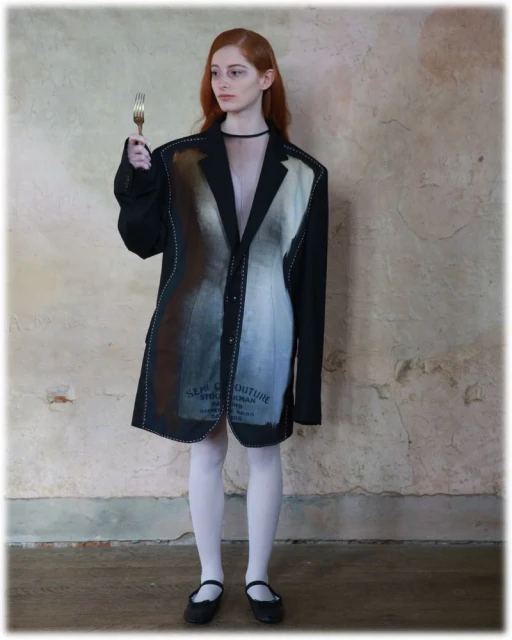 Red-haired model in black couture blazer dress holds fork against distressed wall. Fashion forward portrait showcases unique garment.