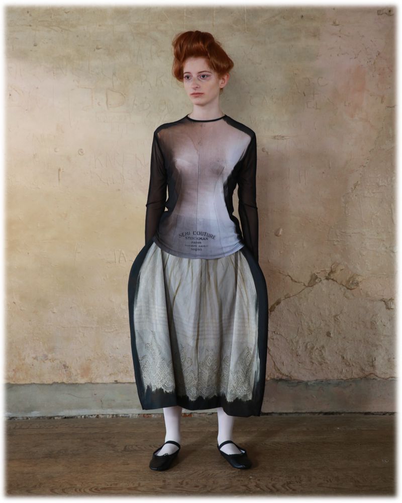 Adult female fashion model wearing a sheer black long-sleeved top, a patterned skirt, white stockings, and black shoes, in a studio.