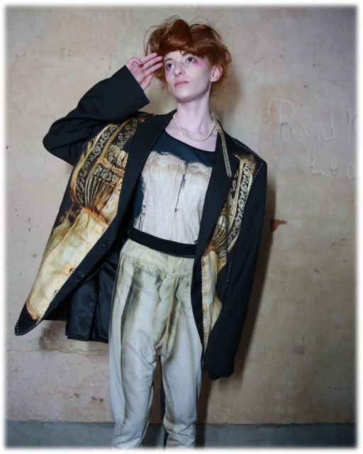 Androgynous person in a black blazer with a graphic print, paired with light-colored pants, poses indoors against a textured wall.