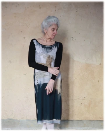 Older woman with silver hair wearing a printed long-sleeve midi dress standing against a textured neutral wall