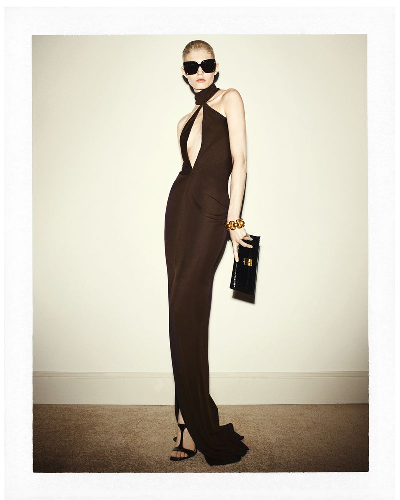 Glamorous fashion shot: Woman in a brown halter gown with sunglasses and a black clutch poses against a neutral backdrop in a studio setting.