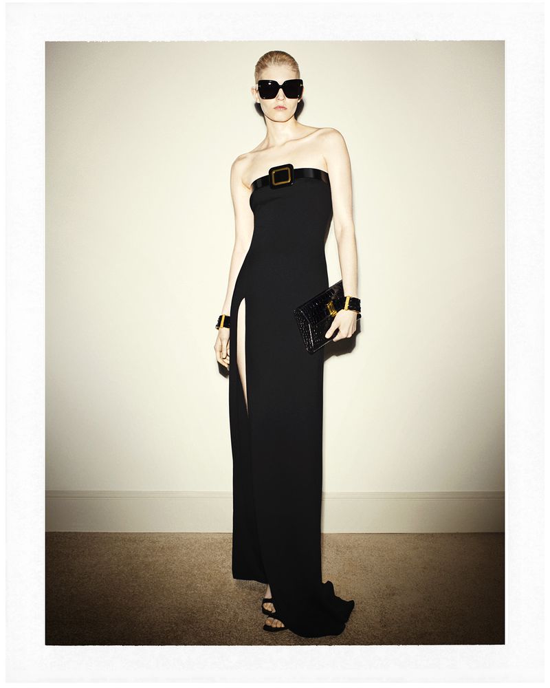 Elegant blonde woman in a black strapless dress with a thigh-high slit, sunglasses, and black clutch poses in front of a plain wall.