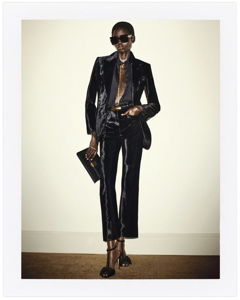 A black woman strikes a fashion pose indoors, wearing a black velvet suit, dark sunglasses, and heels, while holding a black clutch.