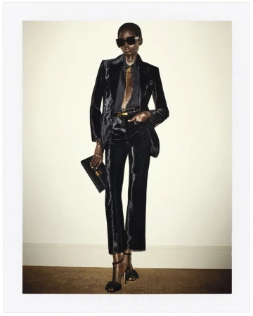 A black woman strikes a fashion pose indoors, wearing a black velvet suit, dark sunglasses, and heels, while holding a black clutch.