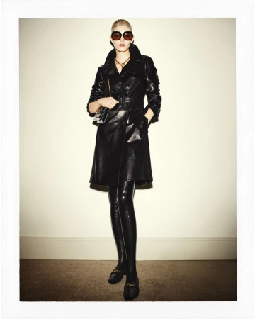 Full-length shot of a woman in a black double-breasted leather coat, thigh-high boots, and sunglasses, holding a small bag against a neutral backdrop.