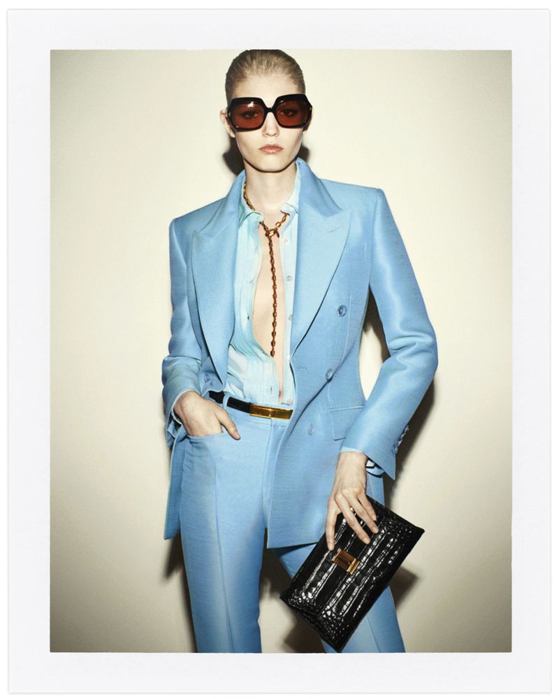 An adult female fashion model poses in a light-blue suit with a black crocodile-pattern clutch, wearing sunglasses in a studio setting.