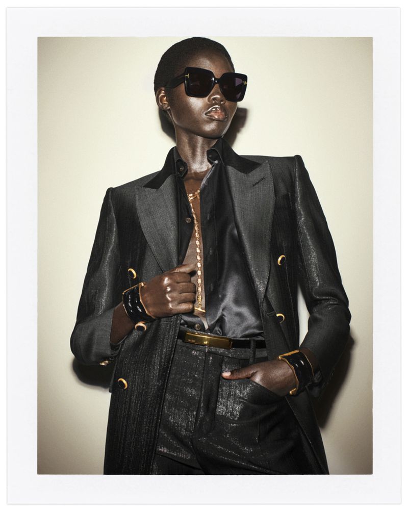 Fashion photo of Black female model in a dark suit with a black shirt, accessorized with gold jewelry and sunglasses, set against a plain background.